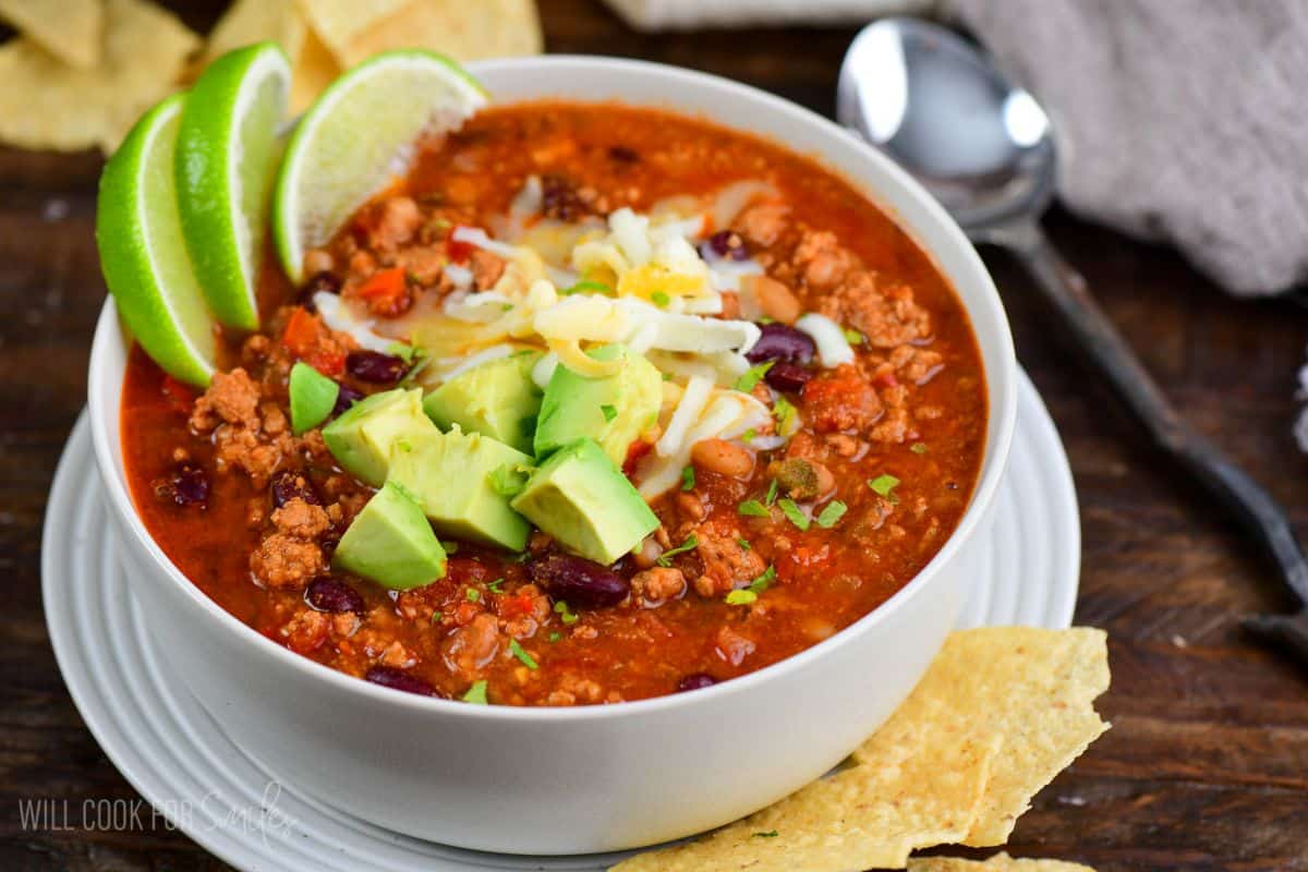 turkey chili in a bowl topped with cheese, avocado, lime wedges, and cilantro.
