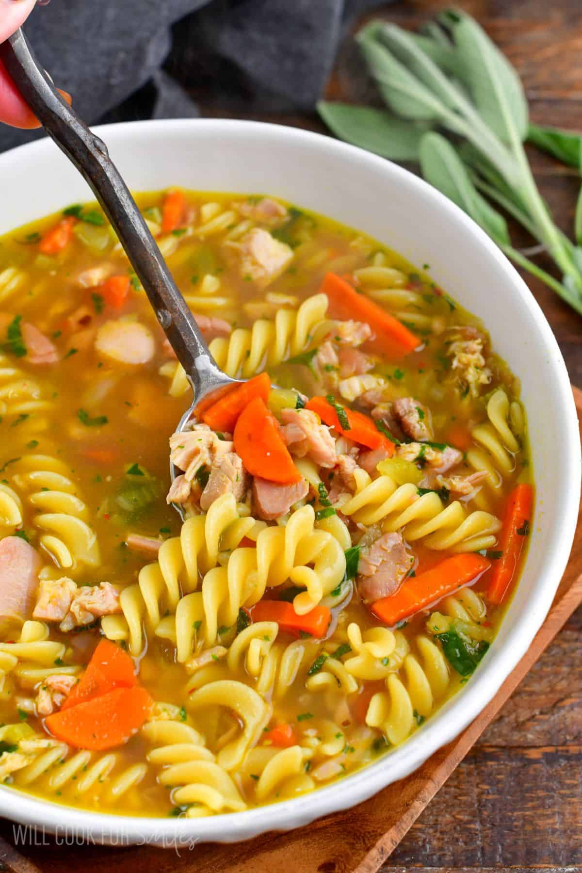 spooning out some comforting and rich turkey noodle soup from a white bowl.