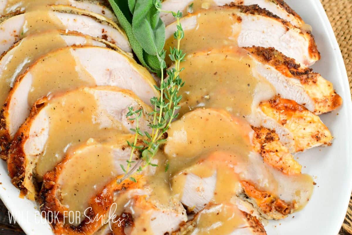 Sliced roasted turkey breast topped with golden gravy and herbs, served on a holiday white platter for Thanksgiving dinner.