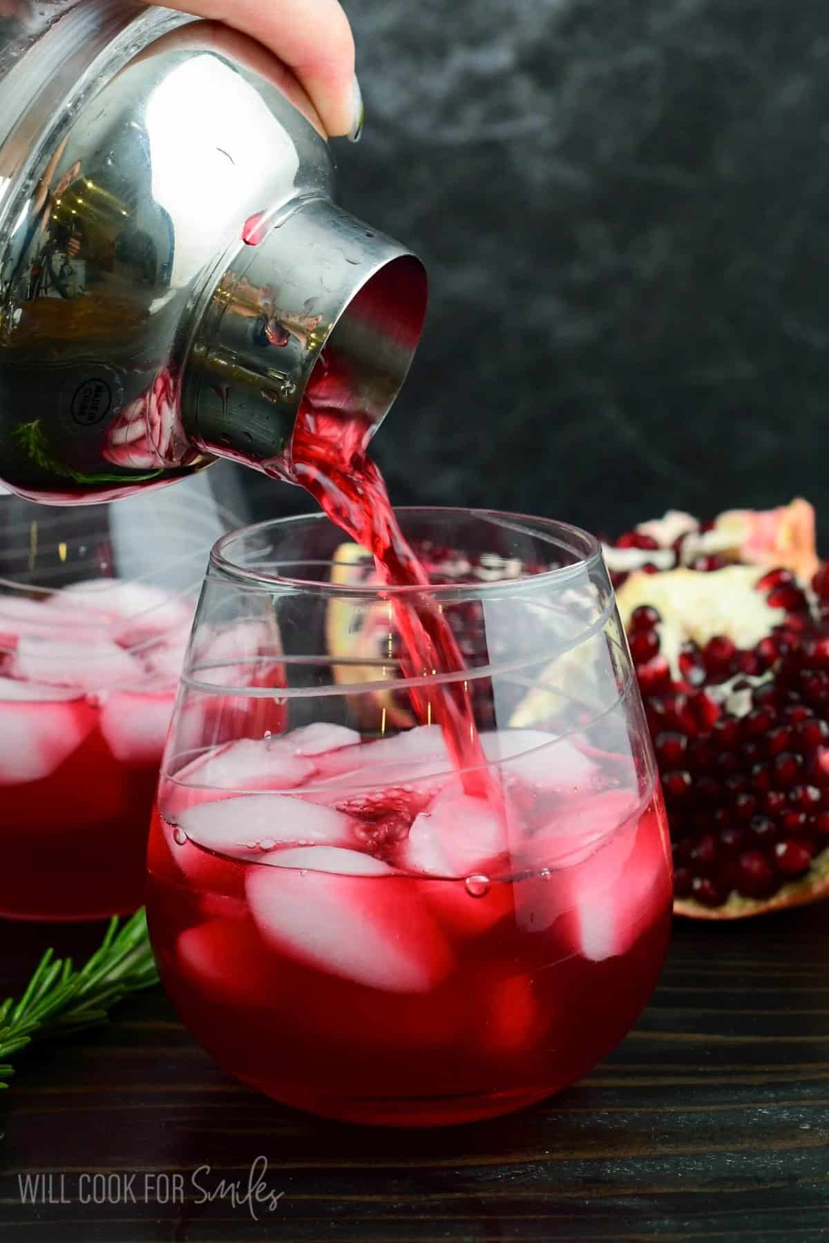 pouring the shaker gin cocktail into the clear glass next to rosemary sprig and open pomegranate.