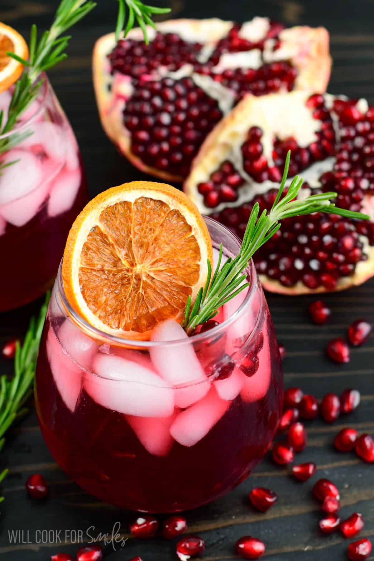 two glasses of pomegranate gin cocktail garnished with orange and rosemary and pomegranate seeds around.