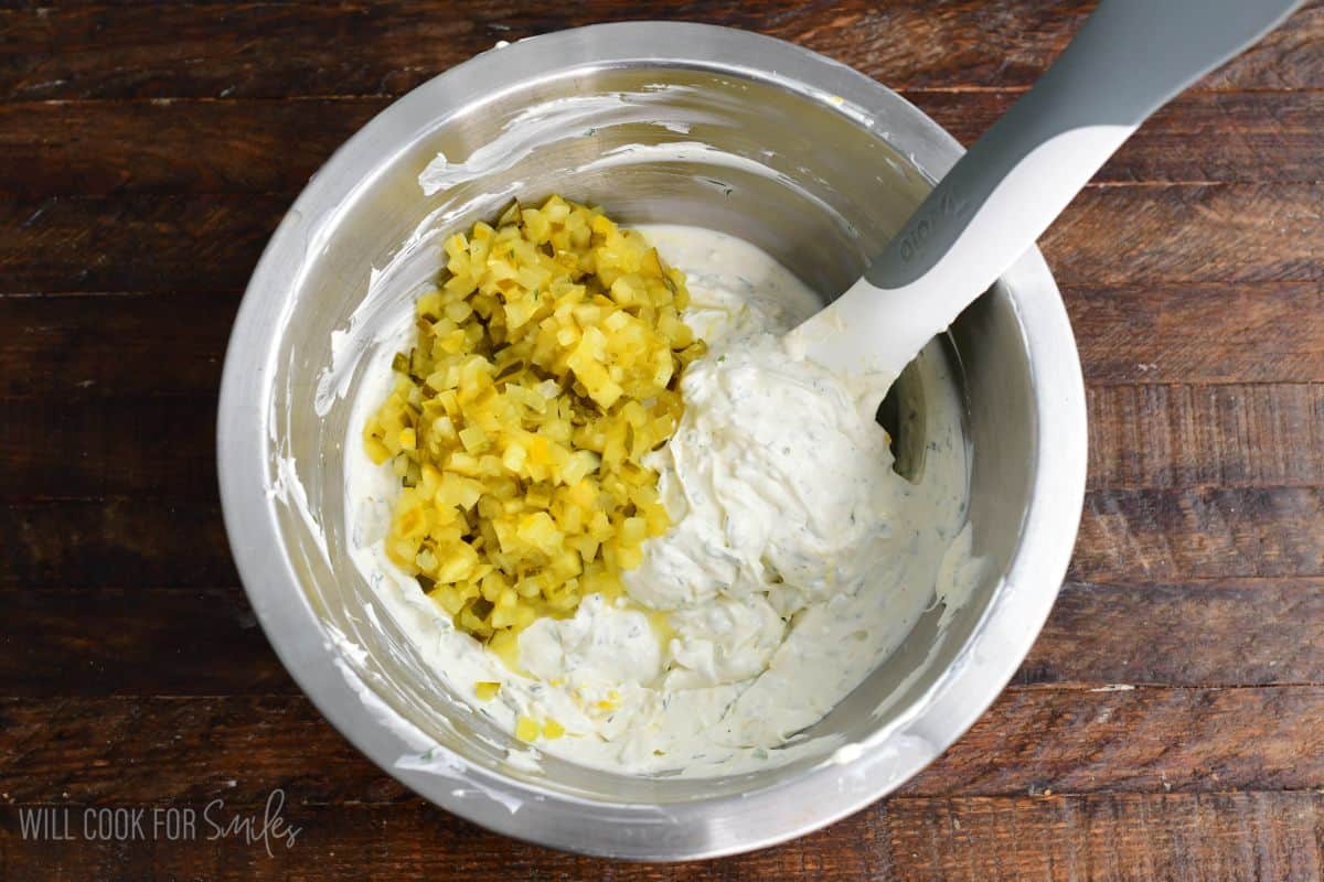 Adding diced pickles to a metal bowl on a wood surface,