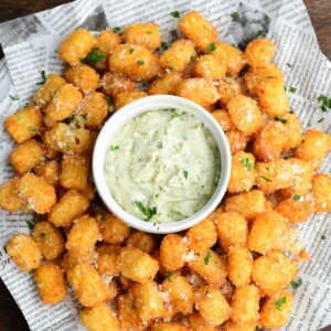 Truffle tater tots on a lined tray with a garlic herb aioli in a bowl in the middle on a wood surface.