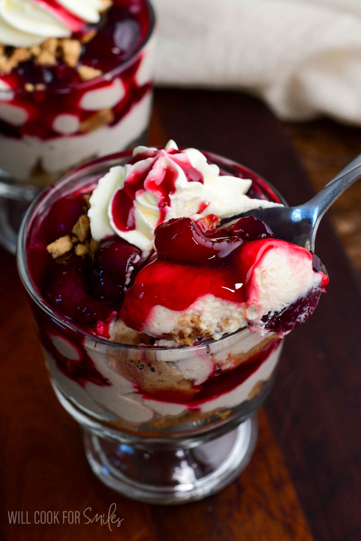 No-bake cherry cheesecake parfait topped with whipped cream and cherry sauce as a spoon scoops into the creamy layers.