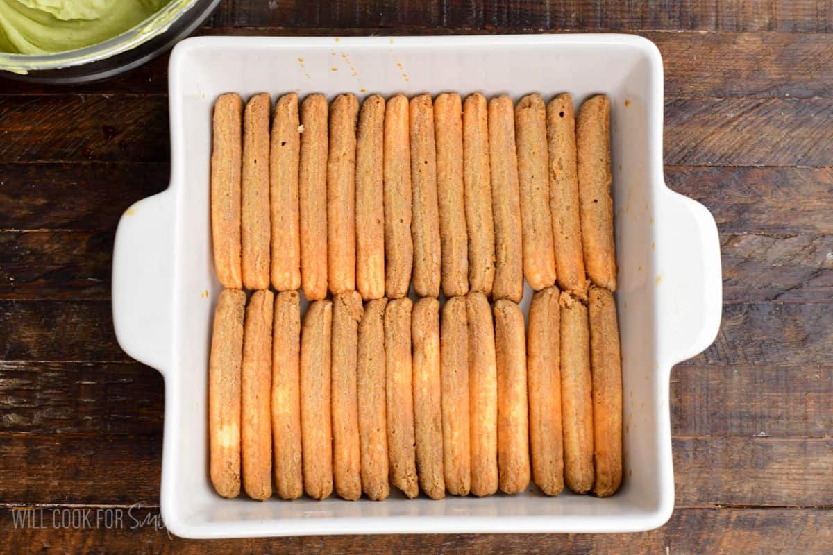 Layer of soaked ladyfinger cookies arranged neatly in a white baking dish, ready for pistachio tiramisu assembly.