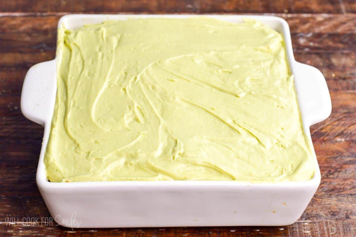 Final layer of creamy pistachio mascarpone mixture spread smoothly over tiramisu in a white baking dish before chilling.