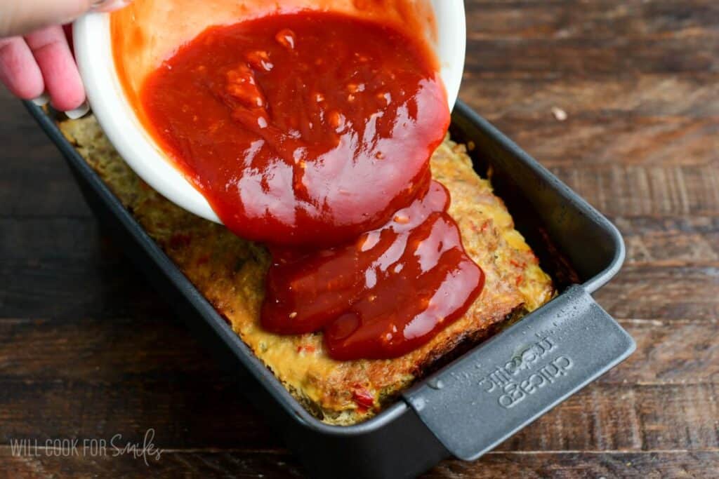 Ketchup glaze being poured over baked turkey meatloaf in a loaf pan, coating the top before returning it to the oven.