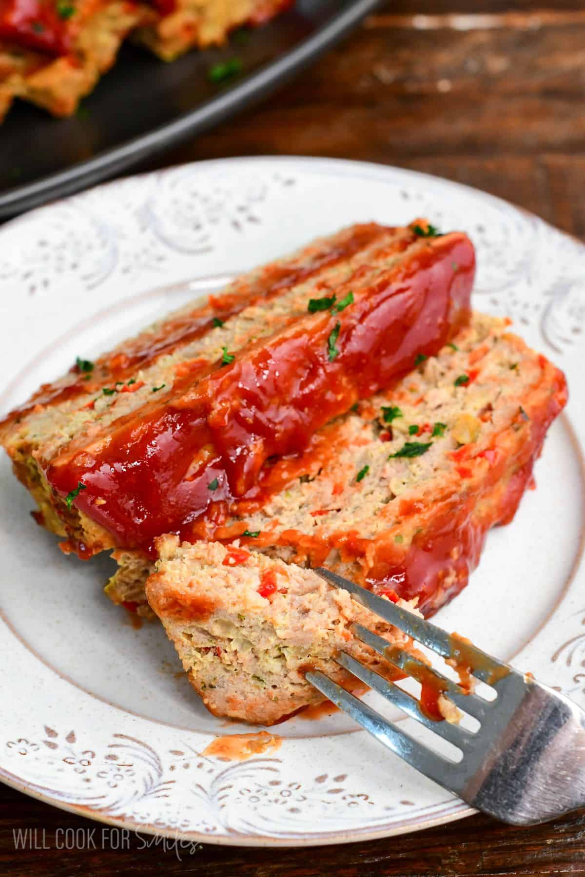 Two slices of turkey meatloaf on a plate with ketchup glaze and parsley, with a fork cutting into a tender bite.